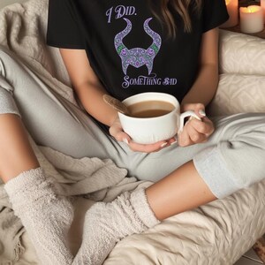 May include: Black t-shirt with the text "I Did. Something Bad" and a purple and green horned design. The person is holding a white mug with a brown liquid. The person is wearing gray sweatpants and fuzzy socks.