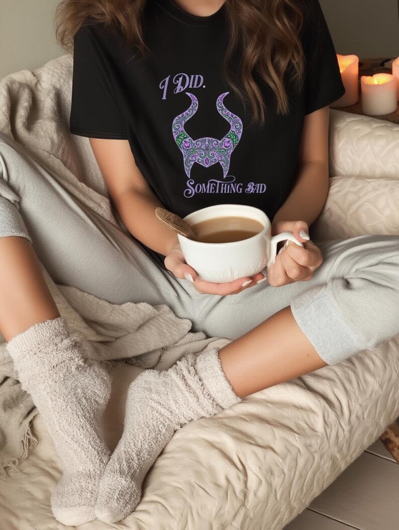 May include: Black t-shirt with the text "I Did. Something Bad" and a purple and green horned design. The person is holding a white mug with a brown liquid. The person is wearing gray sweatpants and fuzzy socks.