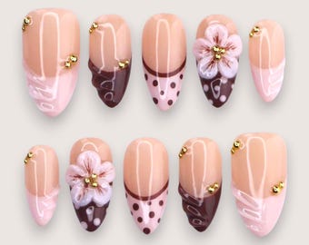 May include: A set of ten artificial nails in various designs. Some nails feature a nude base with a pink tip, others have a dark brown accent with gold embellishments. One nail has a 3D flower.