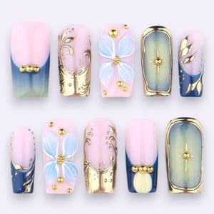 Peut inclure: Un ensemble de dix faux ongles présentant un mélange de motifs, incluant des dégradés rose clair et bleu, des volutes florales métalliques dorées, des pétales de fleurs bleu clair en relief avec des clous dorés et des étoiles dorées.