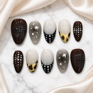 Könnte beinhalten: Ein Set aus zehn mandelförmigen Press-on-Nägeln mit verschiedenen Designs, darunter dunkelbraun mit schwarzen Punktverläufen, grau mit silbernen Metallkugeln, weiße French-Tips mit schwarzen Punkten und gelb mit schwarzen Leopardenmustern.