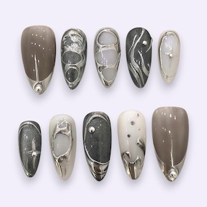Può includere: Un set di dieci unghie finte a mandorla con design assortiti. La collezione presenta basi color tortora lucido, grigio antracite glitterato e bianco latte, arricchite da rilievi metallici argentati che ricordano il metallo liquido.