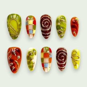 Op de afbeelding: Een set van tien kunstnagels in diverse ontwerpen. De nagels hebben een reeks kleuren, waaronder groen, rood, oranje en geel, met gouden draadaccenten en geometrische patronen. Sommige nagels hebben een spiraalontwerp.