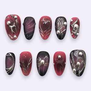 Puede incluir: Un juego de diez uñas postizas con un degradado de rojo intenso y negro. Los diseños incluyen líneas metálicas plateadas en relieve, motivos de corazones y lazos, y pequeñas esferas plateadas. Algunas uñas tienen un acabado liso, mientras que otras presentan texturas onduladas y sutiles destellos brillantes.