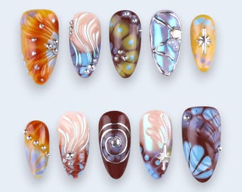 May include: A collection of ten artificial nails with various designs. The nails feature a range of colors, including orange, brown, blue, and pink, with decorative elements like silver studs and swirling patterns. Some nails have star-shaped accents.