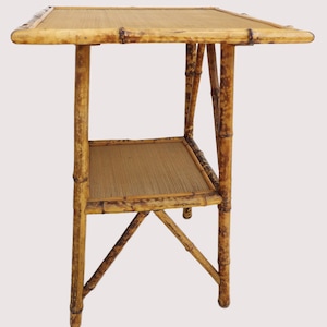 May include: A vintage bamboo side table with a square top and a lower shelf. The table has a natural, light brown color with darker brown accents. The woven top and shelf add texture to the design. The table is approximately 24 inches tall.