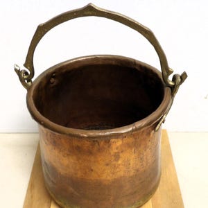 May include: A copper bucket with a brass handle and accents. The bucket has a rounded shape and a dark interior. The handle is arched and attached to the bucket with decorative brass fittings. The bucket sits on a wooden surface.