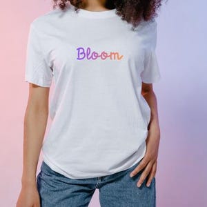 以下が含まれることがあります： 紫からオレンジへのグラデーションで「Bloom」という文字が入った白いTシャツ。青いデニムジーンズと合わせています。背景はピンクと紫のグラデーションです。