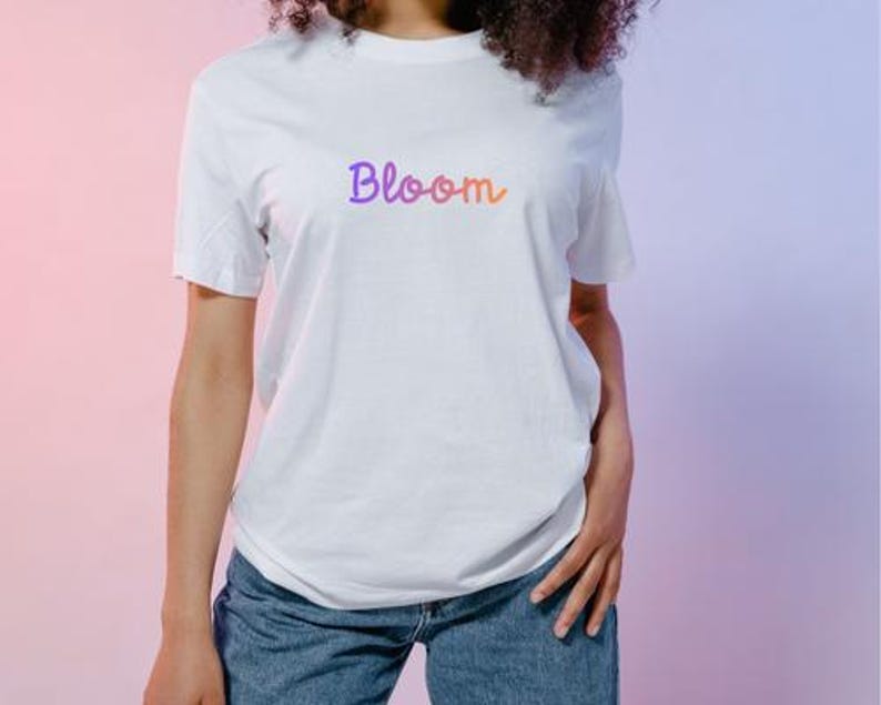 以下が含まれることがあります： 紫からオレンジへのグラデーションで「Bloom」という文字が入った白いTシャツ。青いデニムジーンズと合わせています。背景はピンクと紫のグラデーションです。