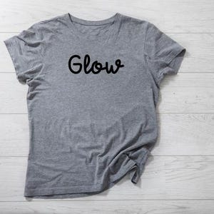 以下が含まれることがあります： 胸に黒い文字で「Glow」と書かれたヘザーグレーのTシャツ。半袖のトップスは柔らかい生地でできています。Tシャツは明るい木製の表面に置かれています。