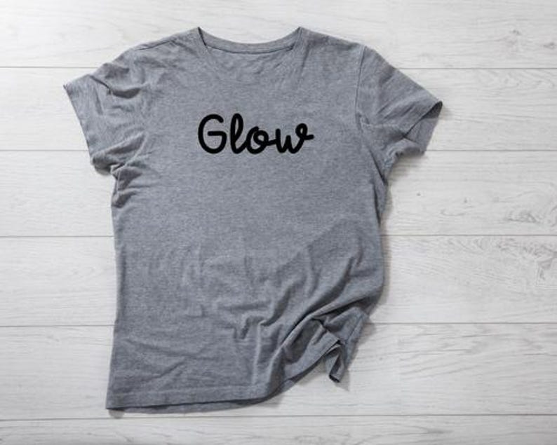 以下が含まれることがあります： 胸に黒い文字で「Glow」と書かれたヘザーグレーのTシャツ。半袖のトップスは柔らかい生地でできています。Tシャツは明るい木製の表面に置かれています。