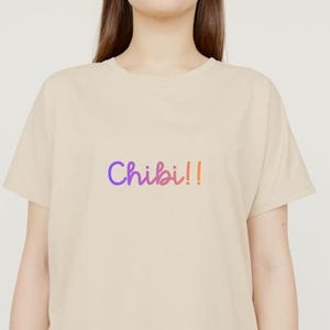 以下が含まれることがあります： 紫からピンクのグラデーションで「Chibi!!」という文字が入ったベージュのTシャツ。Tシャツは半袖で、クルーネックです。テキストは手書き風です。