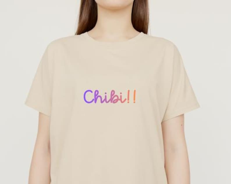 以下が含まれることがあります： 紫からピンクのグラデーションで「Chibi!!」という文字が入ったベージュのTシャツ。Tシャツは半袖で、クルーネックです。テキストは手書き風です。