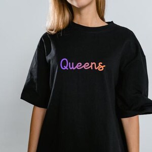 以下が含まれることがあります： 紫からオレンジへのグラデーションで「Queens」という文字が入った黒いTシャツ。Tシャツは半袖で、クルーネックです。テキストは筆記体です。