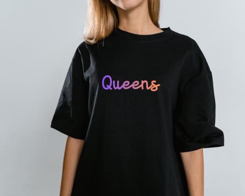以下が含まれることがあります： 紫からオレンジへのグラデーションで「Queens」という文字が入った黒いTシャツ。Tシャツは半袖で、クルーネックです。テキストは筆記体です。