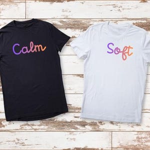 以下が含まれることがあります： 木製の表面に、黒と白の2枚のTシャツが写っています。黒いTシャツには、紫、オレンジ、黄色のグラデーションで「Calm」という文字が書かれています。白いTシャツには、同様のグラデーションで「Soft」という文字が書かれています。