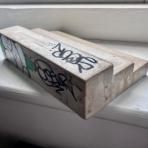 Pode incluir: Uma borda de concreto bege com design escalonado, apresentando letras de grafite pretas nas faces frontal e lateral. O objeto possui uma superfície áspera e texturizada, com formato retangular e um nível elevado com marcas de tinta.