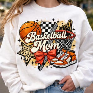 Könnte beinhalten: Weißes Sweatshirt mit einem Basketball-Motiv. Das Design zeigt einen Basketball, einen Korb, Turnschuhe und den Text "Basketball Mom". Akzente sind ein Stern, eine Schleife und ein Schaumstoff-Finger.