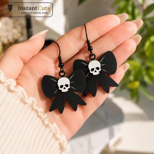 May include: A pair of black drop earrings featuring a bow shape with a white skull center. The earrings have black metal hooks and are designed with a flat, matte finish. The design includes the text InstantCuts Laser Cut Digital Files.