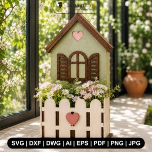 Könnte beinhalten: Ein dekorativer, hausförmiger Pflanzkübel aus Holz mit hellgrüner Fassade, dunkelbraunem Dach und einem weißen Lattenzaun als Basis. Ein kleines rosa Herz ist auf dem Dach und dem Zaun angebracht. Der Kübel ist mit weißen und rosa Blumen gefüllt.