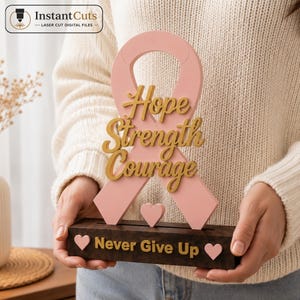 May include: A light pink awareness ribbon cutout featuring the gold text Hope Strength Courage, mounted on a dark brown rectangular base with the gold text Never Give Up and two small pink hearts. The design includes a small pink heart cutout.