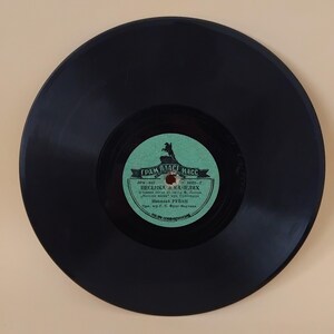 Pode incluir: Um disco de vinil preto com um rótulo verde e branco. O rótulo apresenta texto em língua estrangeira e um gráfico de um cavalo e cavaleiro. O disco está centrado numa superfície bege clara.