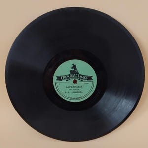 Pode incluir: Um disco de vinil preto com um rótulo verde e branco. O rótulo apresenta texto em cirílico e uma ilustração de um cavalo e um cavaleiro. O disco está centrado numa superfície bege clara.