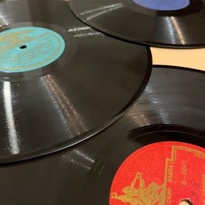 Pode incluir: Close-up de vários discos de vinil pretos com rótulos coloridos. Os rótulos são de várias cores, incluindo vermelho, azul e turquesa. Os discos estão dispostos em um padrão ligeiramente sobreposto, mostrando sua forma circular e superfície brilhante.