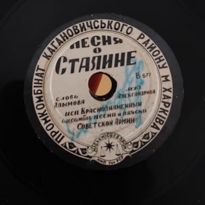 Pode incluir: Um disco de vinil preto vintage com um rótulo branco e azul. O rótulo apresenta texto cirílico, um orifício central e um design circular. O disco é uma peça da história da música.