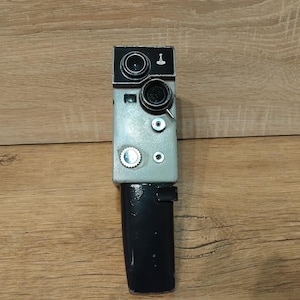 May include: A vintage, handheld film camera with a gray body, black top and bottom, and multiple lenses and dials. The camera has a rectangular shape and is positioned vertically against a wooden background. The camera's design suggests it is from a bygone era.