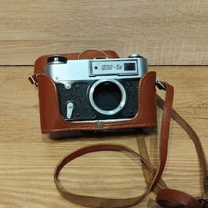 May include: A vintage ФЭД-5B camera in a brown leather case with a matching strap. The camera body is silver and black, with a prominent lens and the model name printed on the top. The camera is resting on a wooden surface.