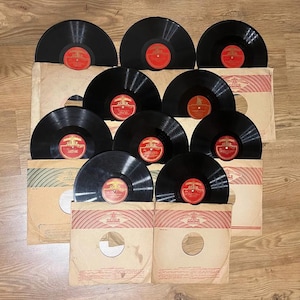 Pode incluir: Uma coleção de discos de vinil vintage pretos com rótulos vermelhos e dourados. Estão guardados em capas de papel bege com detalhes vermelhos e brancos, dispostos sobre uma superfície de madeira.