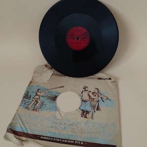 Pode incluir: Um disco de vinil vintage com uma superfície azul escura e um rótulo vermelho. O disco repousa sobre uma capa de papel desgastada com ilustrações de figuras em trajes tradicionais e um caçador. A capa tem texto em língua estrangeira.