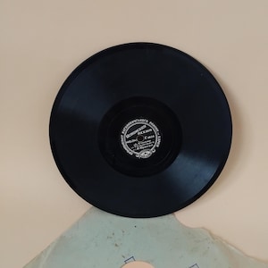 Pode incluir: Um disco de vinil preto com um rótulo com texto cirílico e um emblema decorativo. O disco está centrado em uma superfície bege clara, com uma porção de uma capa de papel visível abaixo.