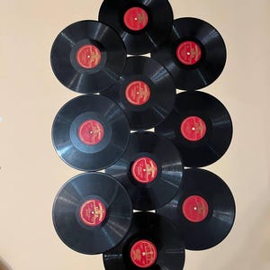Pode incluir: Uma coleção de dez discos de vinil pretos com rótulos vermelhos e dourados. Os discos estão dispostos em forma de leque sobre um fundo neutro. Os rótulos apresentam texto e elementos decorativos.
