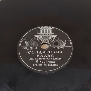 Pode incluir: Um disco de vinil preto vintage com um rótulo cinza e branco. O rótulo apresenta texto cirílico, o título "Soldatskiy Vals" e o logotipo do fabricante. O disco tem um orifício central e ranhuras visíveis.
