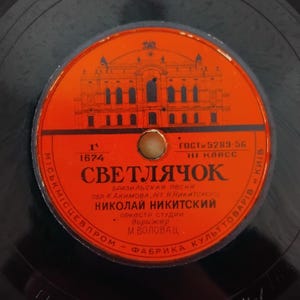 Pode incluir: Close-up de um disco de vinil vintage com um rótulo laranja vibrante. O rótulo apresenta texto cirílico e uma ilustração de um edifício. O disco é preto com um rótulo circular.