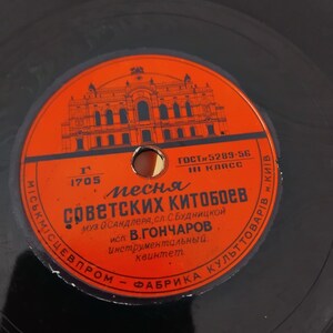 Pode incluir: Um disco de vinil vintage com um rótulo laranja com texto cirílico e uma ilustração de um edifício. O rótulo do disco inclui o texto "ГОСТ 5289-56" e "1705". O disco é preto e parece estar em boas condições.