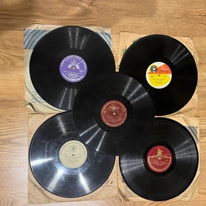 Pode incluir: Cinco discos de vinil vintage com rótulos coloridos e capas de papel. Os discos pretos estão dispostos sobre uma superfície de madeira. Os rótulos apresentam texto e desenhos em várias cores, incluindo vermelho, amarelo e roxo.