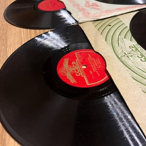 Pode incluir: Vários discos de vinil vintage são exibidos, mostrando suas superfícies pretas e rótulos coloridos. Alguns discos são parcialmente visíveis dentro de suas capas de papel, com rótulos vermelhos e laranja. Os discos estão dispostos em uma superfície de madeira.