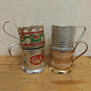 May include: Four vintage metal cup holders with handles. Two are silver-toned, one with a red and gold design, and the other with a celestial pattern. The other two are gold-toned, one with a red and green floral design.