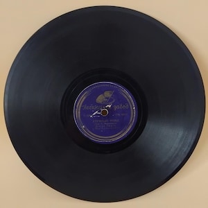 May include: A vintage black vinyl record with a purple and white label. The label features text in a foreign language and a graphic of a hat and a ship. The record is circular and has grooves.