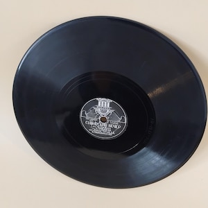 Peut inclure: Un disque vinyle noir avec une étiquette comportant du texte en langue étrangère. Le disque présente des rainures concentriques et une surface brillante. L'étiquette est blanche et grise avec du texte et un logo.