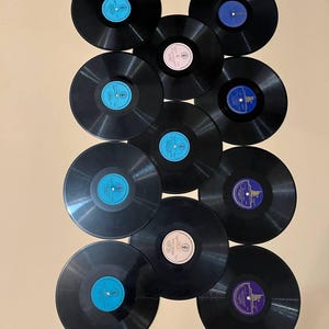 May include: A collection of black vinyl records with colorful labels, including blue, pink, and purple. The records are arranged on a beige background, showcasing their circular shape and vintage aesthetic.