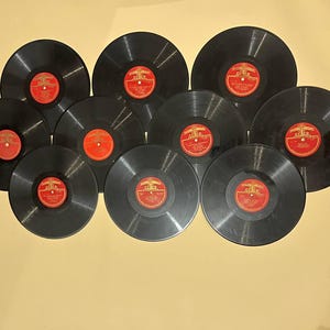 Pode incluir: Uma coleção de dez discos de vinil pretos com rótulos vermelhos e dourados. Os discos estão dispostos em um grupo, mostrando sua forma circular e os detalhes dos rótulos. Os discos são um item de música vintage.