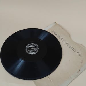 Pode incluir: Um disco de vinil preto com um rótulo branco com texto cirílico, repousando sobre uma capa de papel desgastada. A capa tem texto cirílico impresso. O disco é um item de música vintage.