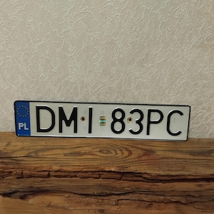 May include: A rectangular Polish license plate with black lettering and numbers on a white background. The plate reads "DM I 83PC" and features a blue EU emblem with the letters "PL". The plate is mounted on a wooden surface.