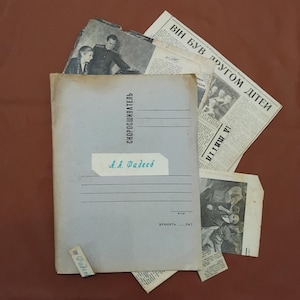 May include: A collection of vintage documents, including a light blue folder with the text "СКОРОСШИВАТЕЛЬ" and "А.А. Фадеев". Several newspaper clippings and a small photograph are scattered around the folder, all on a brown surface.