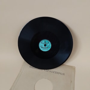 Pode incluir: Um disco de vinil preto com um rótulo turquesa e branco com texto cirílico. O disco é colocado sobre uma superfície bege clara, com uma porção de uma capa de papel visível abaixo. A capa tem texto cirílico.