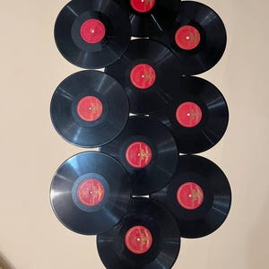 Pode incluir: Uma coleção de dez discos de vinil pretos com rótulos vermelhos. Os discos estão dispostos verticalmente, mostrando sua forma circular e o contraste entre o vinil preto e os rótulos vermelhos.